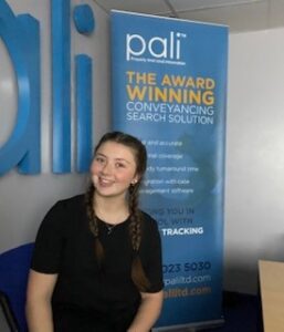 Amber Ritson, a team member from Pali Ltd., smiling and sitting in front of a company banner. The banner displays information about Pali's award-winning conveyancing search solutions. Amber is wearing a black shirt and has her hair styled in braids.